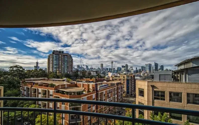 View from a balcony overlooking a city skyline with buildings, trees, and a cloudy blue sky.