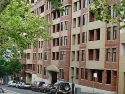 Apartment building with brick facade and tan panels, street lined with cars, trees in foreground.