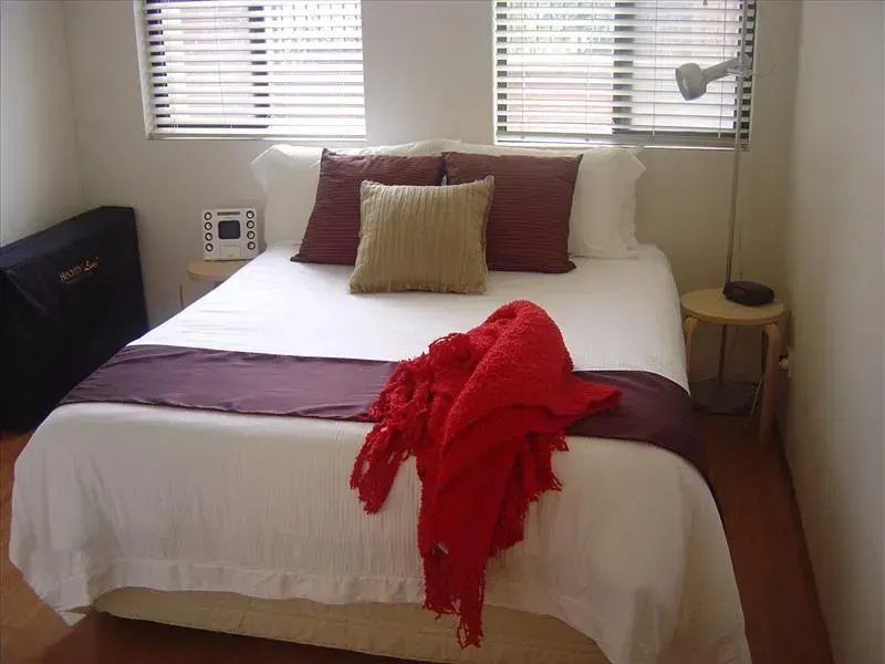 Bedroom with a made bed, white linens, burgundy pillows, and red throw. Windows above.