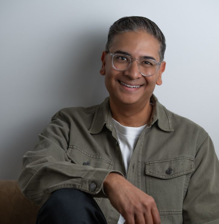 Smiling person wearing glasses and an olive jacket, seated against a light gray background.