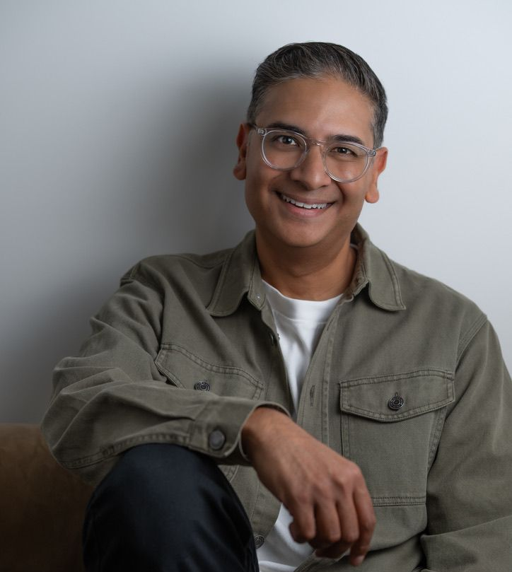 Smiling person with glasses in an olive shirt, seated against a light gray background