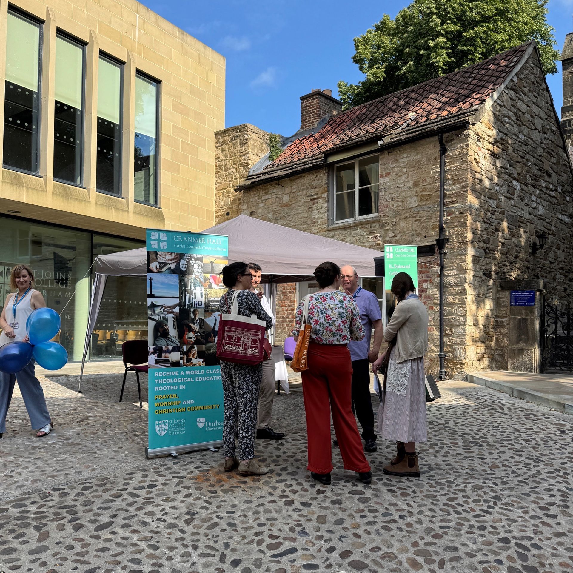 People gathered at an outdoor event beside stone and glass buildings under a canopy.