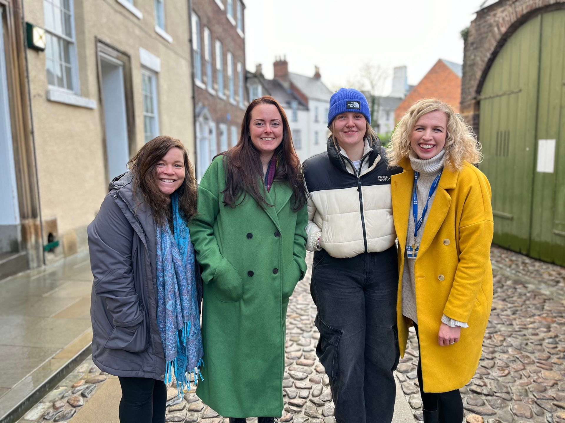 Four people smiling outdoors on a narrow street, wearing colorful coats and standing by old buildings.