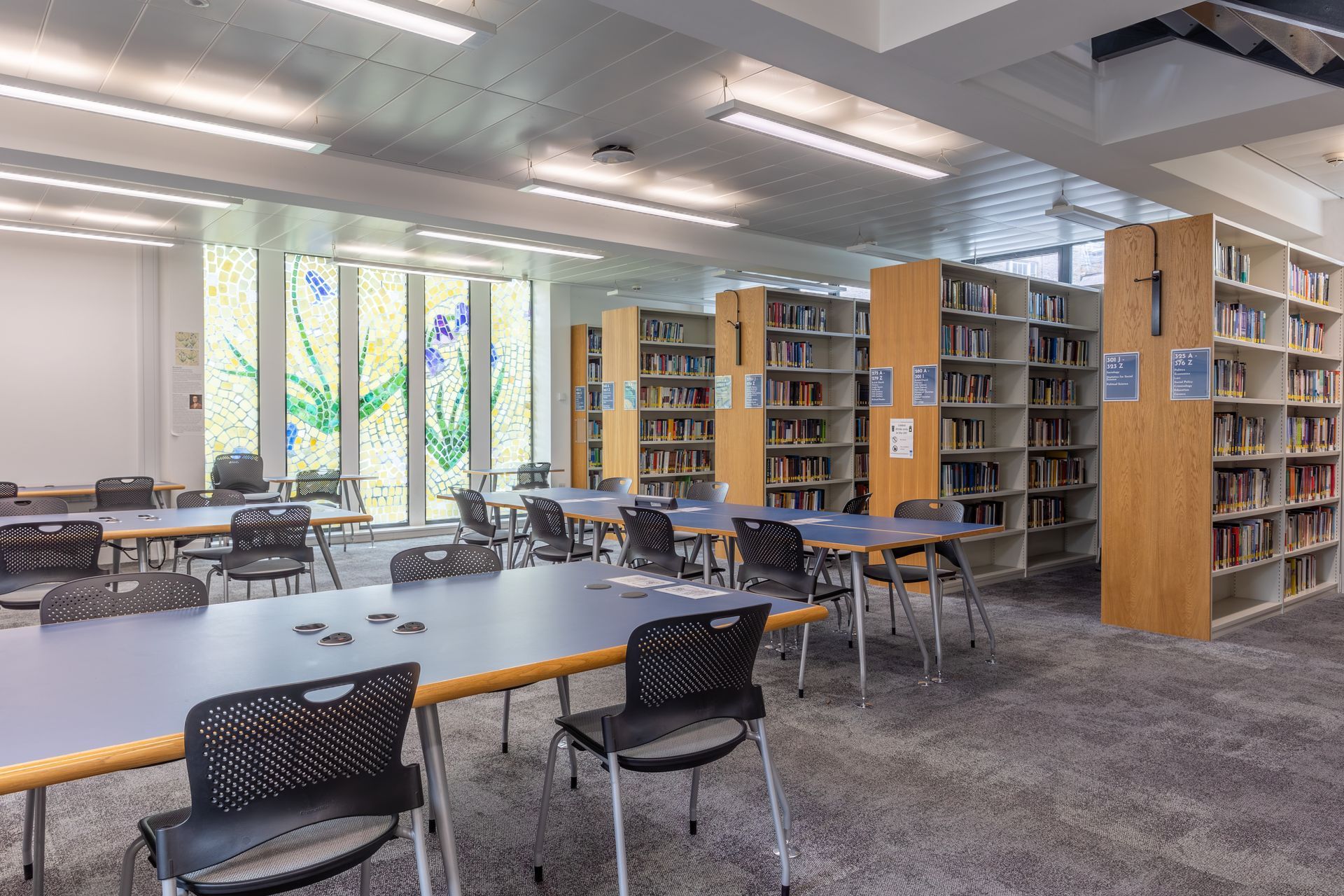 Bright library study room with tables, chairs, bookshelves, and large windows.