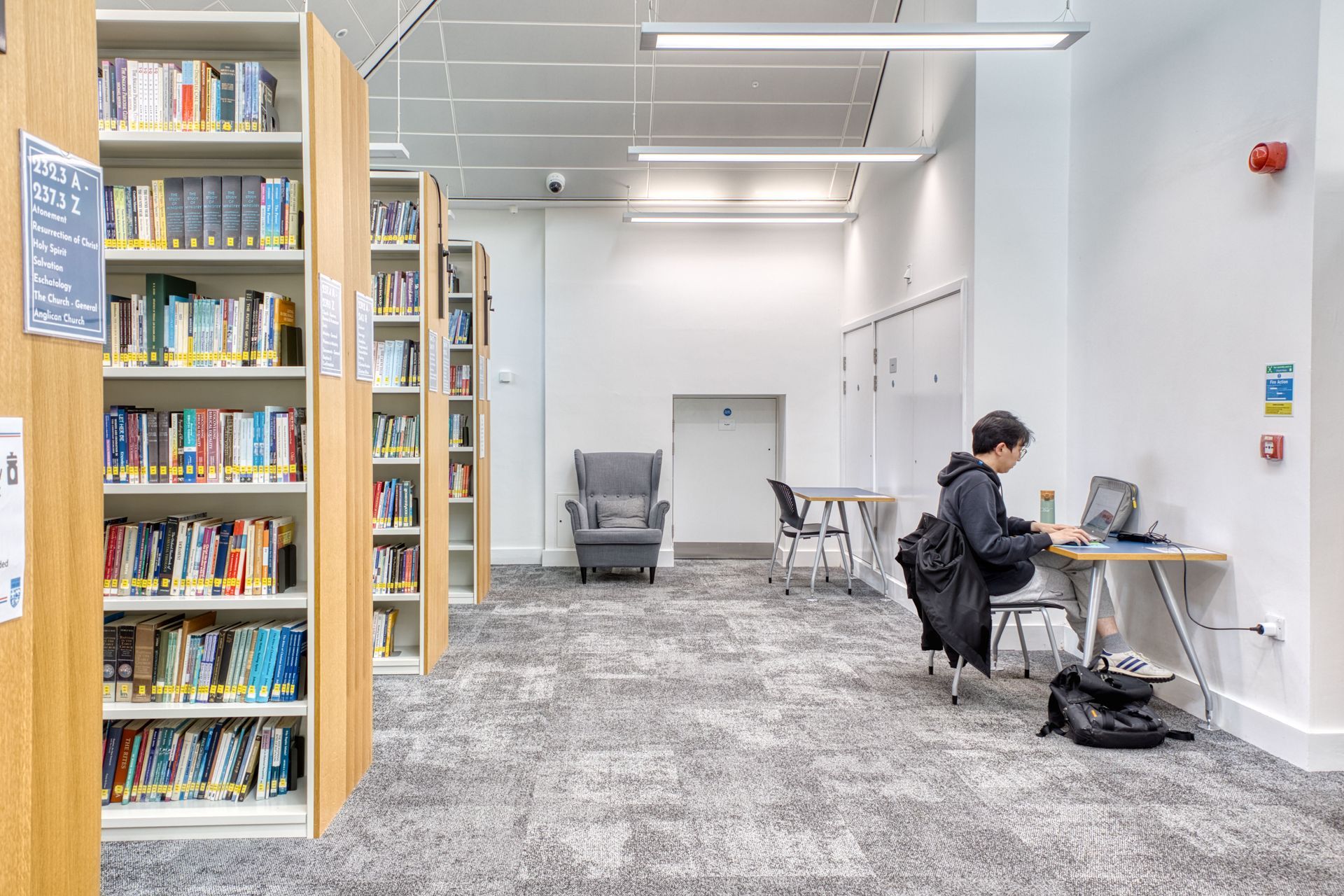 Quiet library study area with bookshelves, tables, and a person working on a laptop.