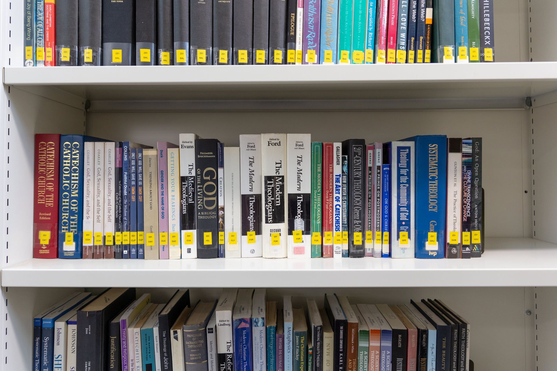 Books arranged on white shelves in a library or study, with colorful spines and labels visible.