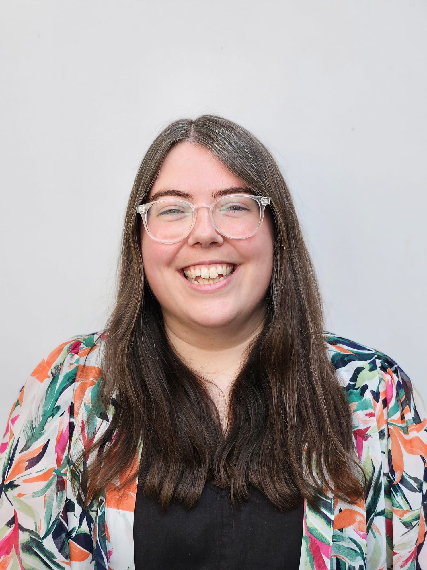 Smiling person with long dark hair and glasses, wearing a colorful patterned top over black against a light background