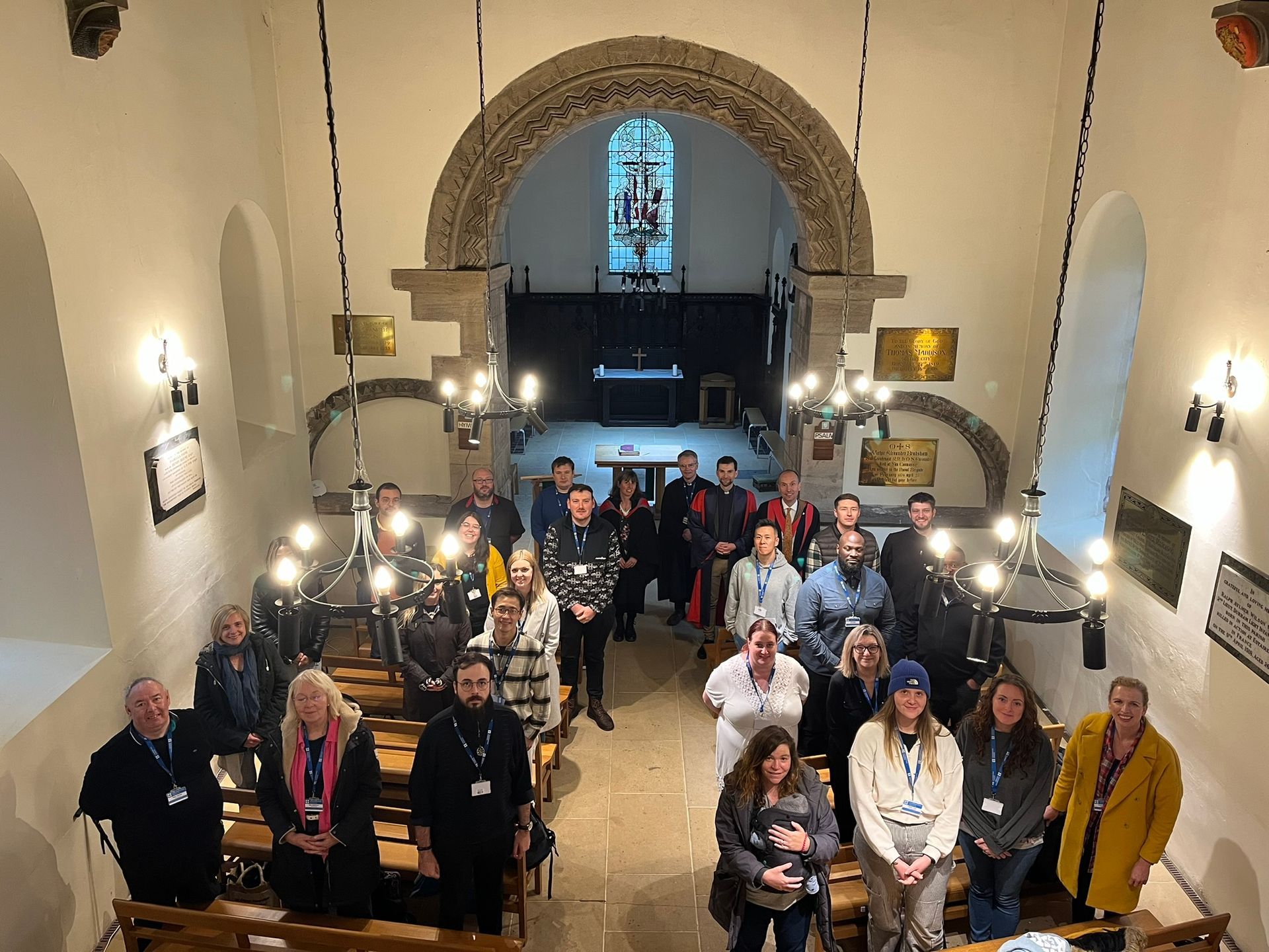 Group of people standing in a church or chapel interior beneath an arched stained-glass window.
