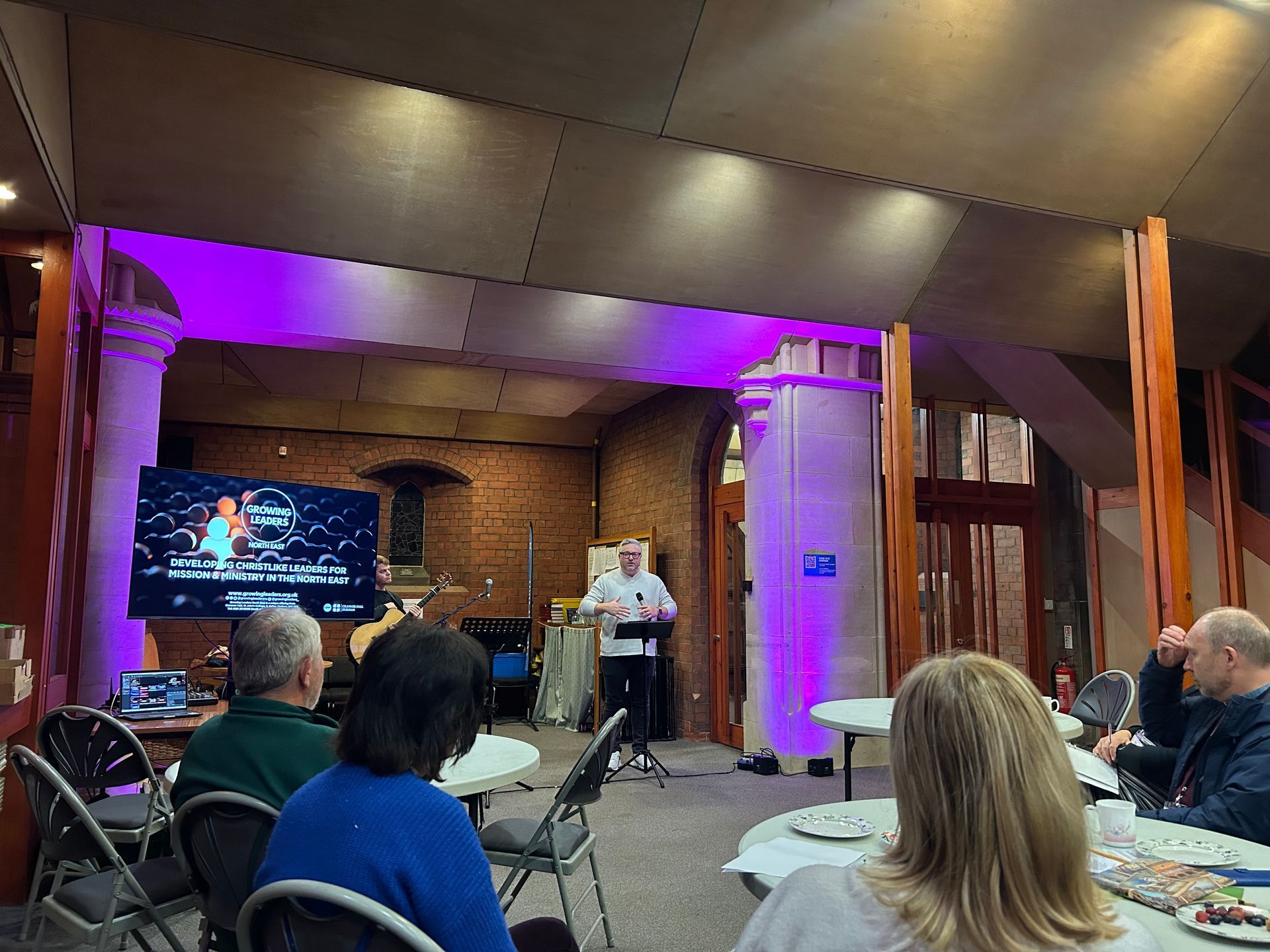 Event presentation in a brick hall with purple lighting, screen on stage, and seated audience.
