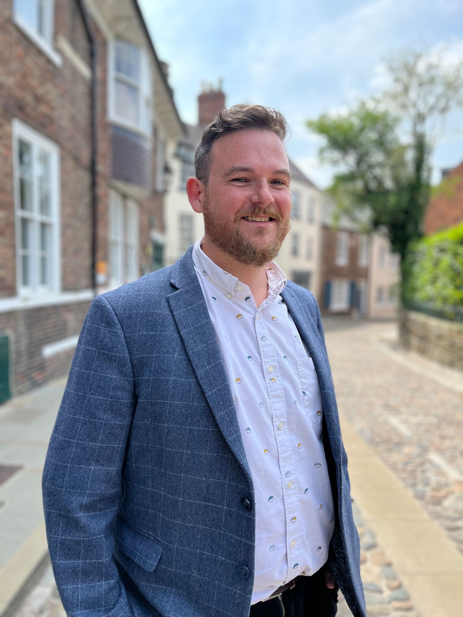 Man in a blue blazer standing outdoors on a sunny street beside brick buildings.