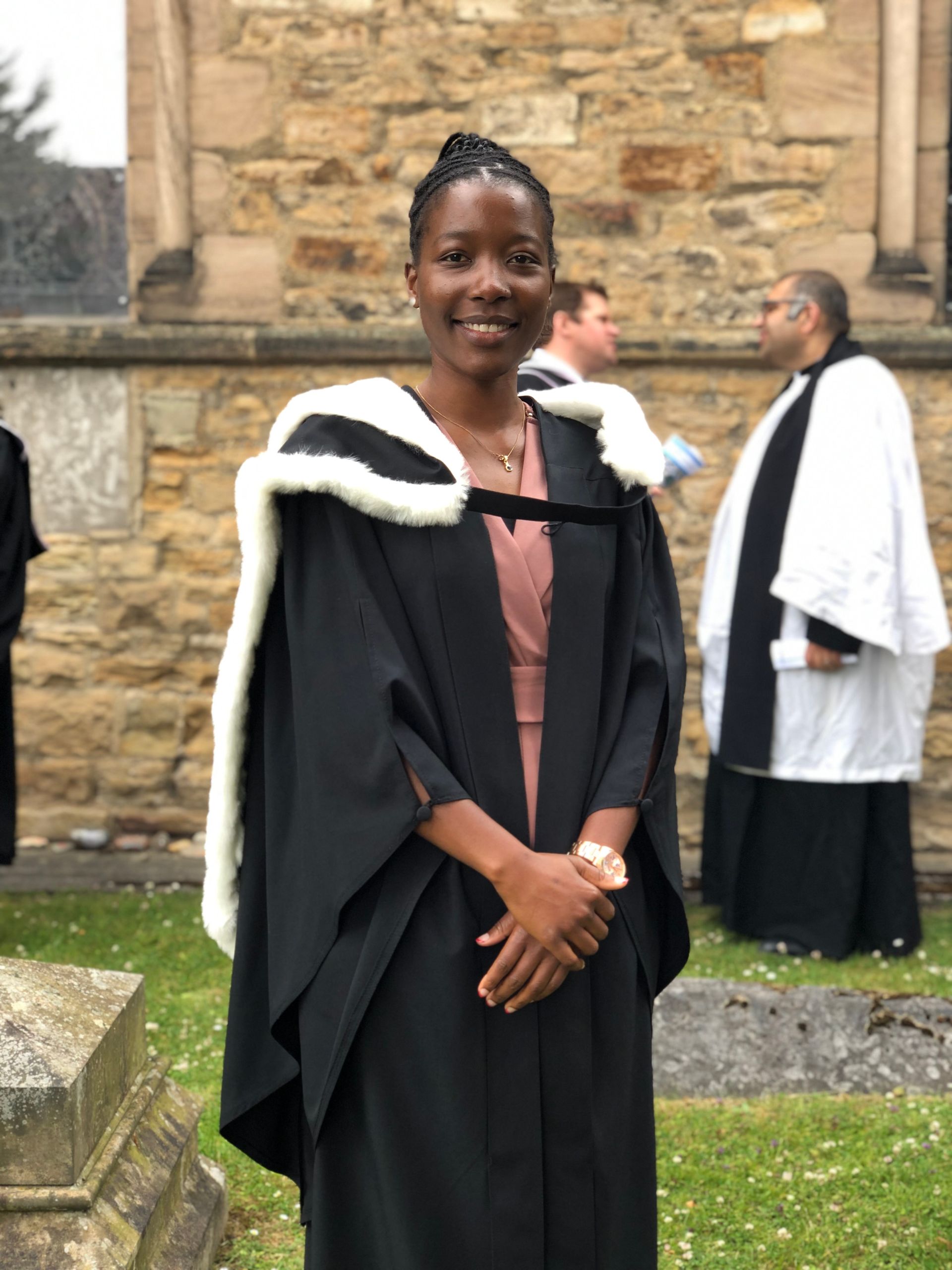 Person in a black graduation gown smiling outdoors in front of a stone building