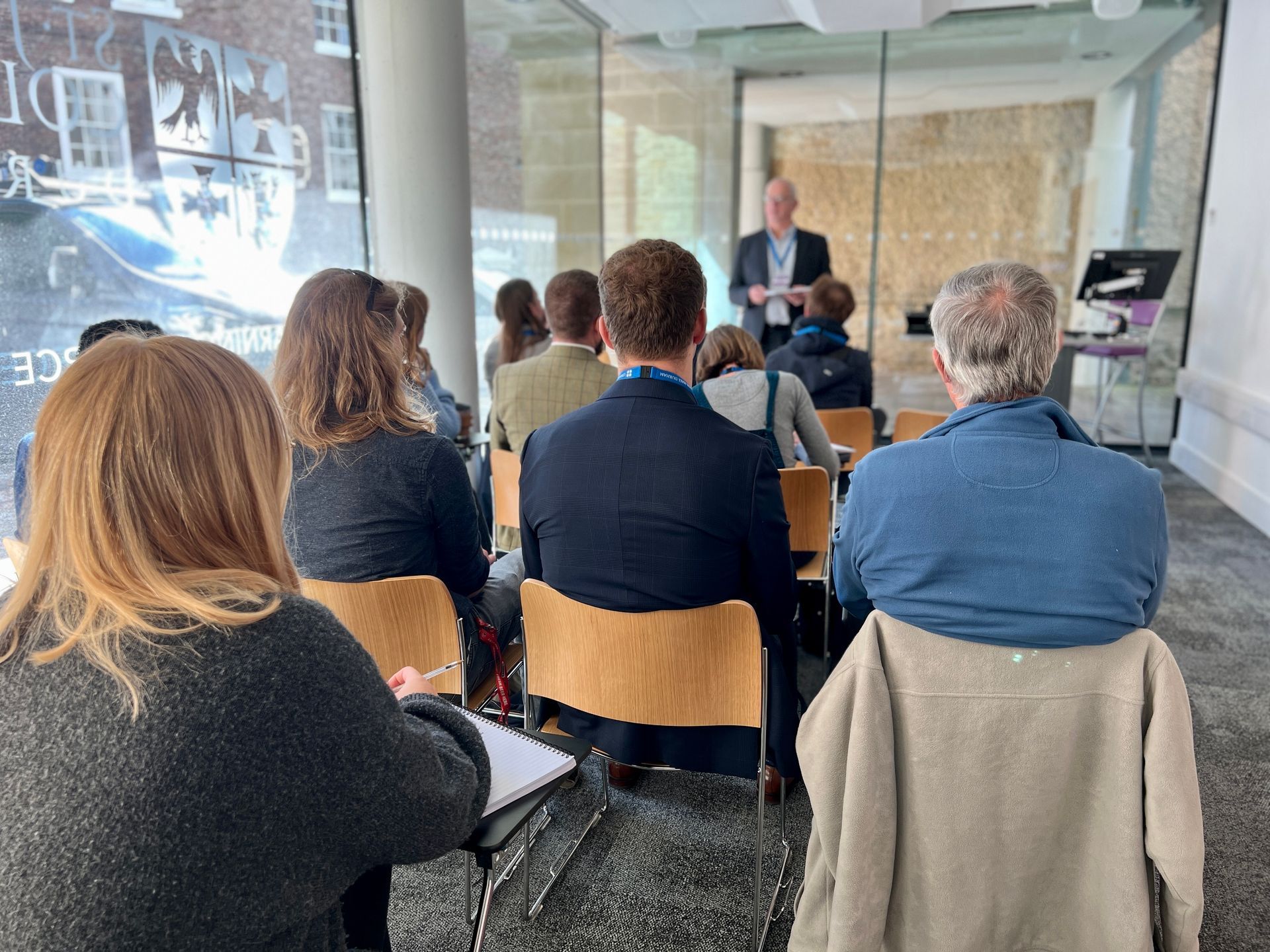 Audience seated in a glass-walled classroom listening to a speaker at the front