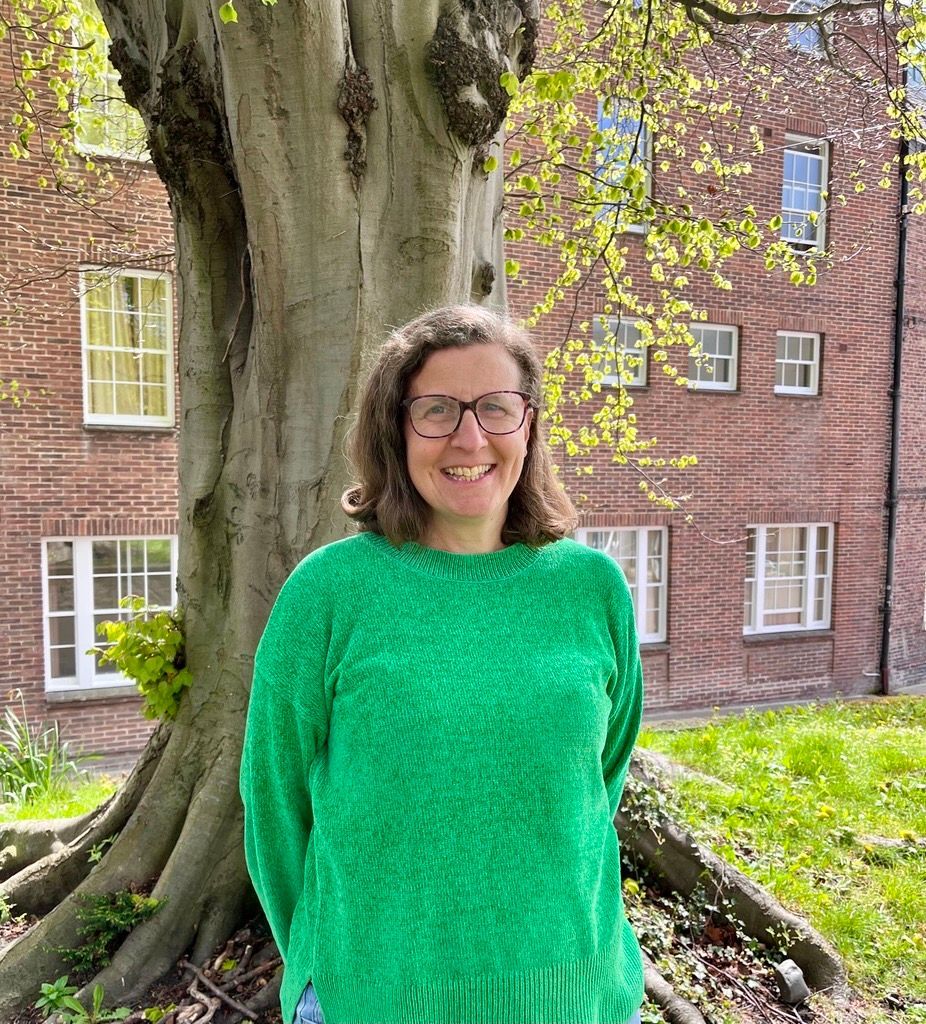 Smiling bald man with glasses and beard in a dark blue shirt, standing outdoors with greenery behind him