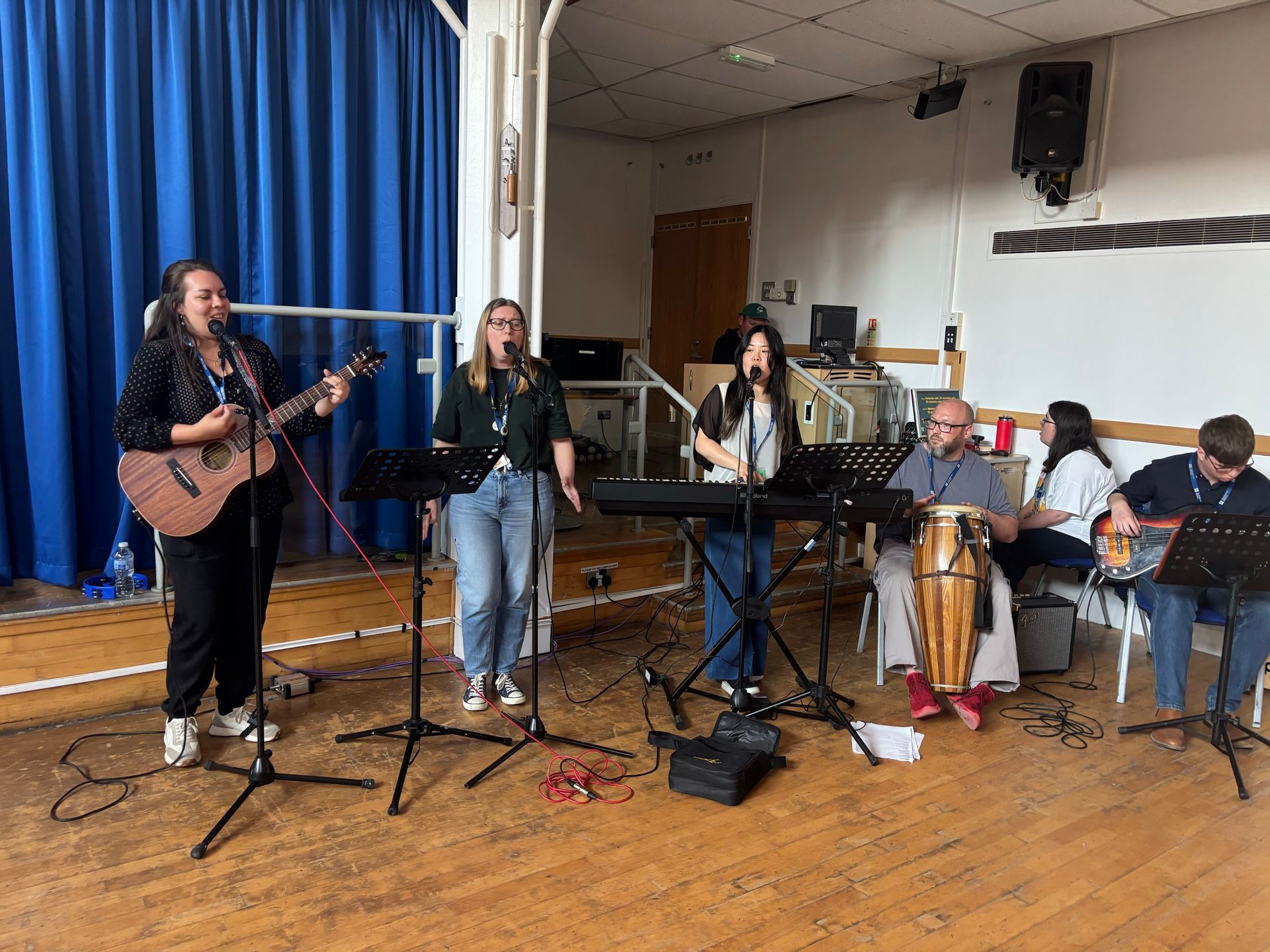 Small band performing on a wooden stage with microphones, guitars, accordion, and blue curtains.