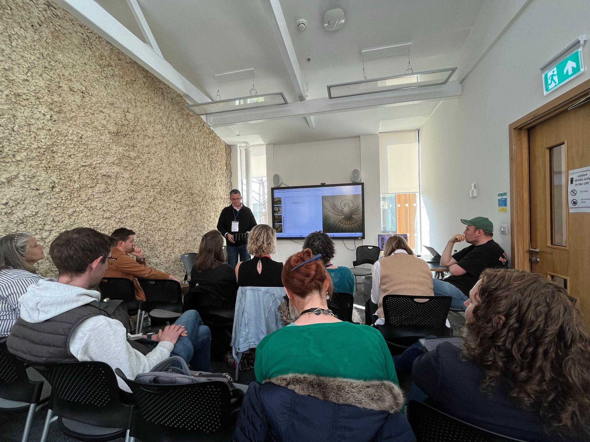 People seated in a small classroom watching a presenter near a screen.