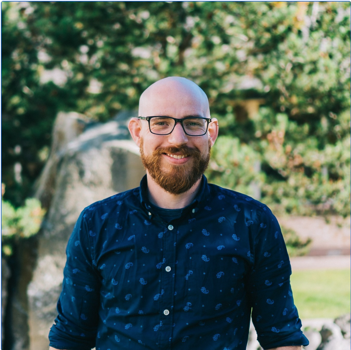 Smiling bald man with glasses and beard in a dark blue shirt, standing outdoors with greenery behind him