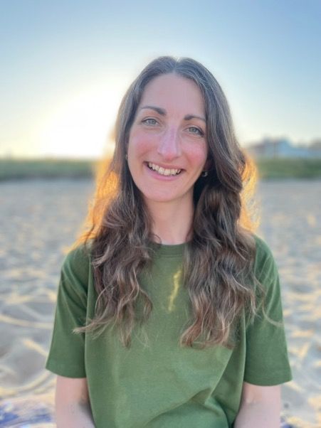 Smiling person in a green shirt standing outdoors on a sandy beach at sunset