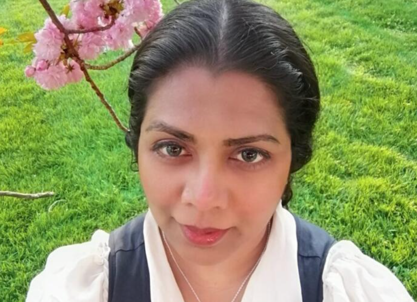 Close-up selfie of a person with a neutral expression in a green garden with pink flowers behind them