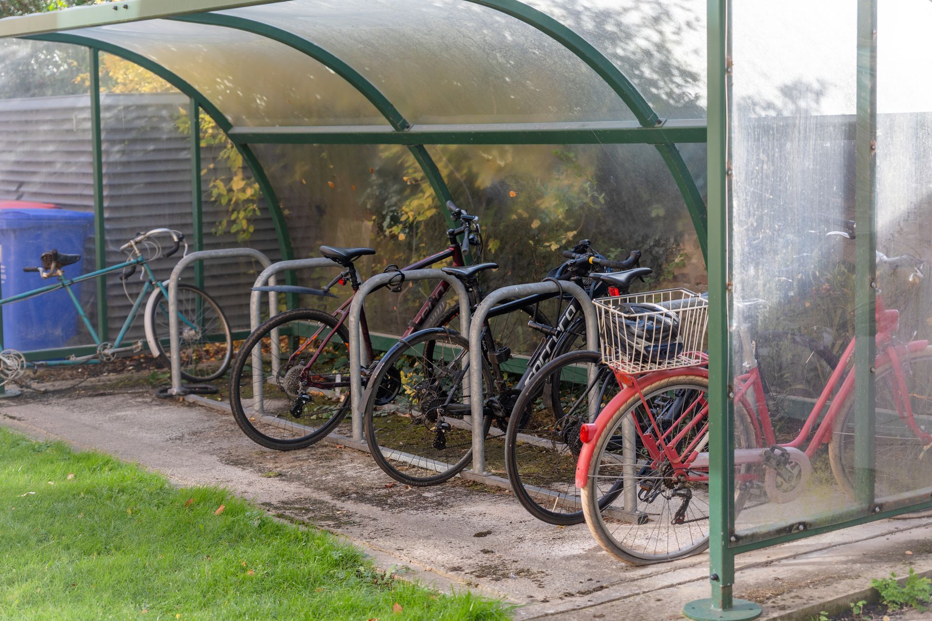 Bicycles parked under a transparent green bike shelter beside a grassy path.