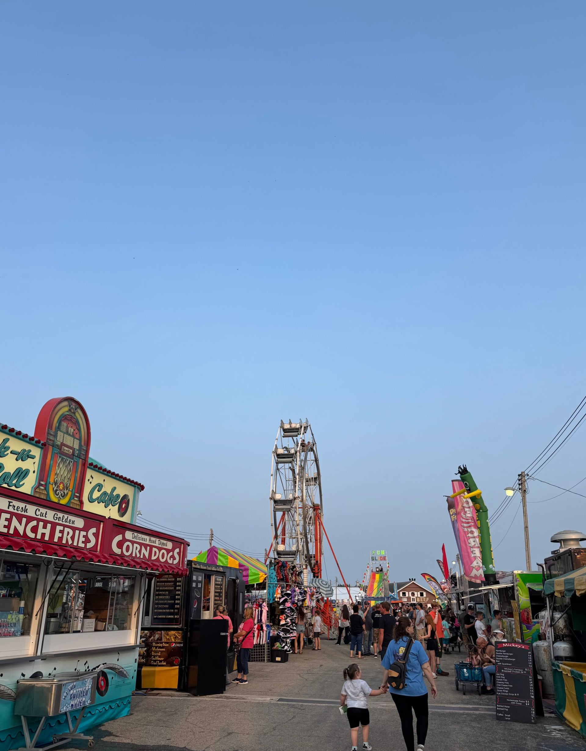 Crowd walking through a sunny outdoor carnival with a Ferris wheel, thrill ride, and food stalls selling corndogs.
