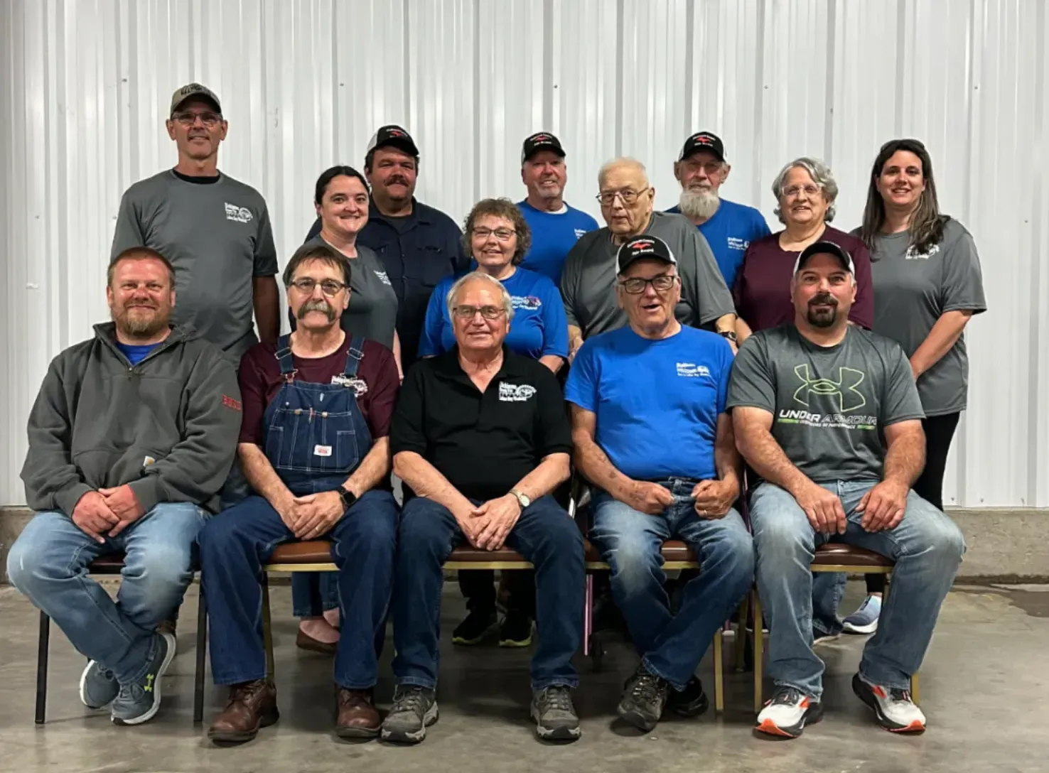 A group of 15 people smiling, posed in two rows in front of a white corrugated wall.