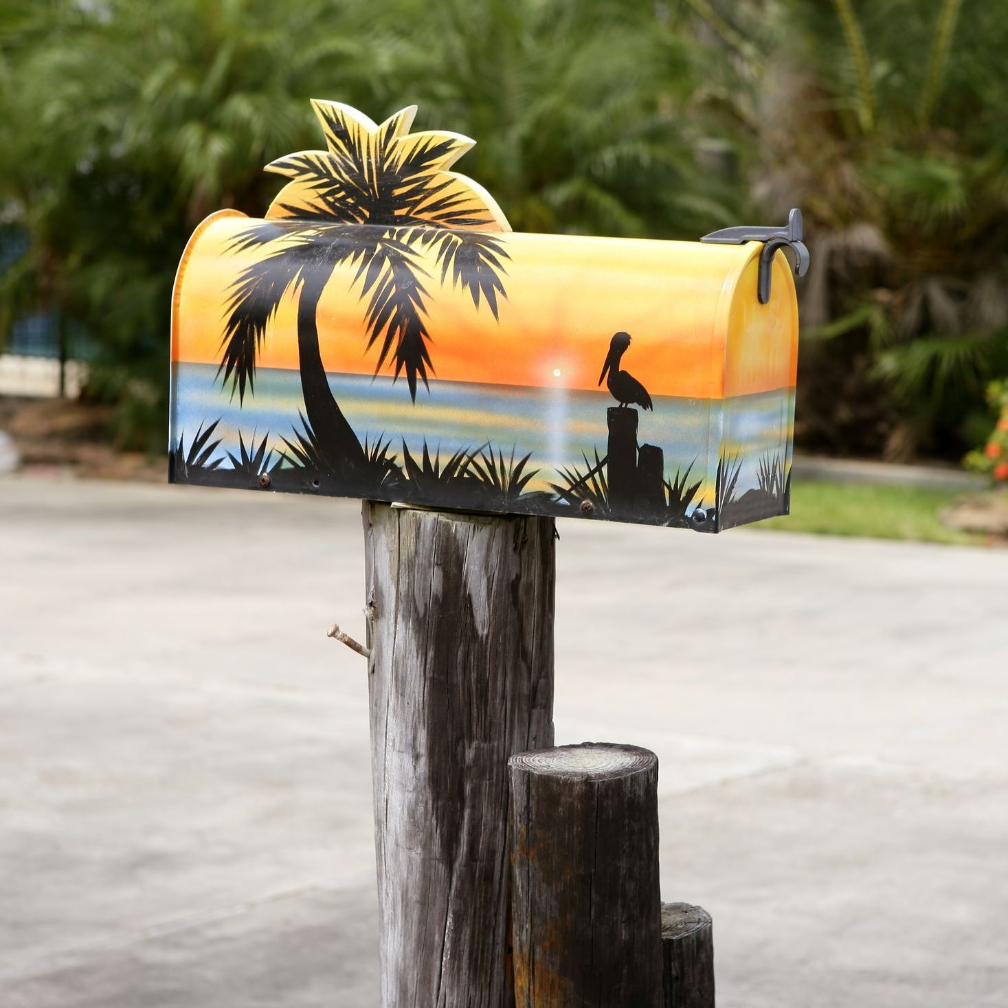 Beach scene mailbox with palm tree silhouette, pelican, and sunset sky, mounted on a weathered post.
