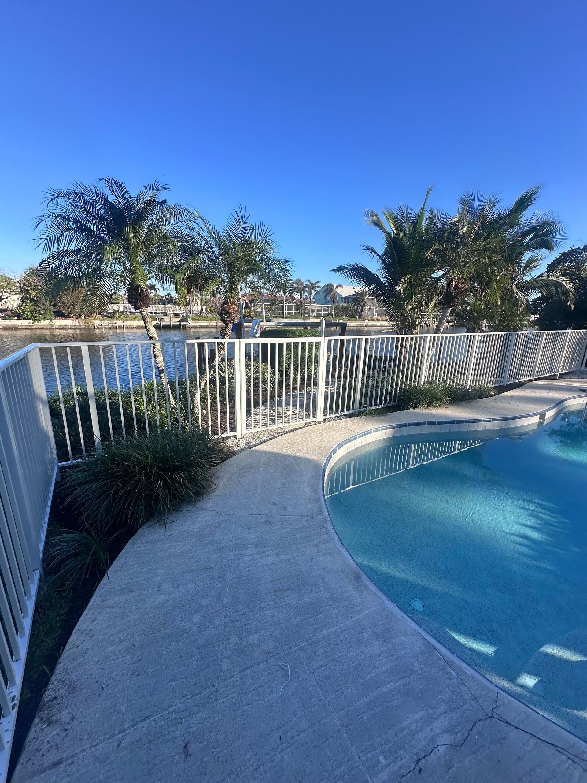 Poolside view: white fence, blue water, and trees under a clear sky.
