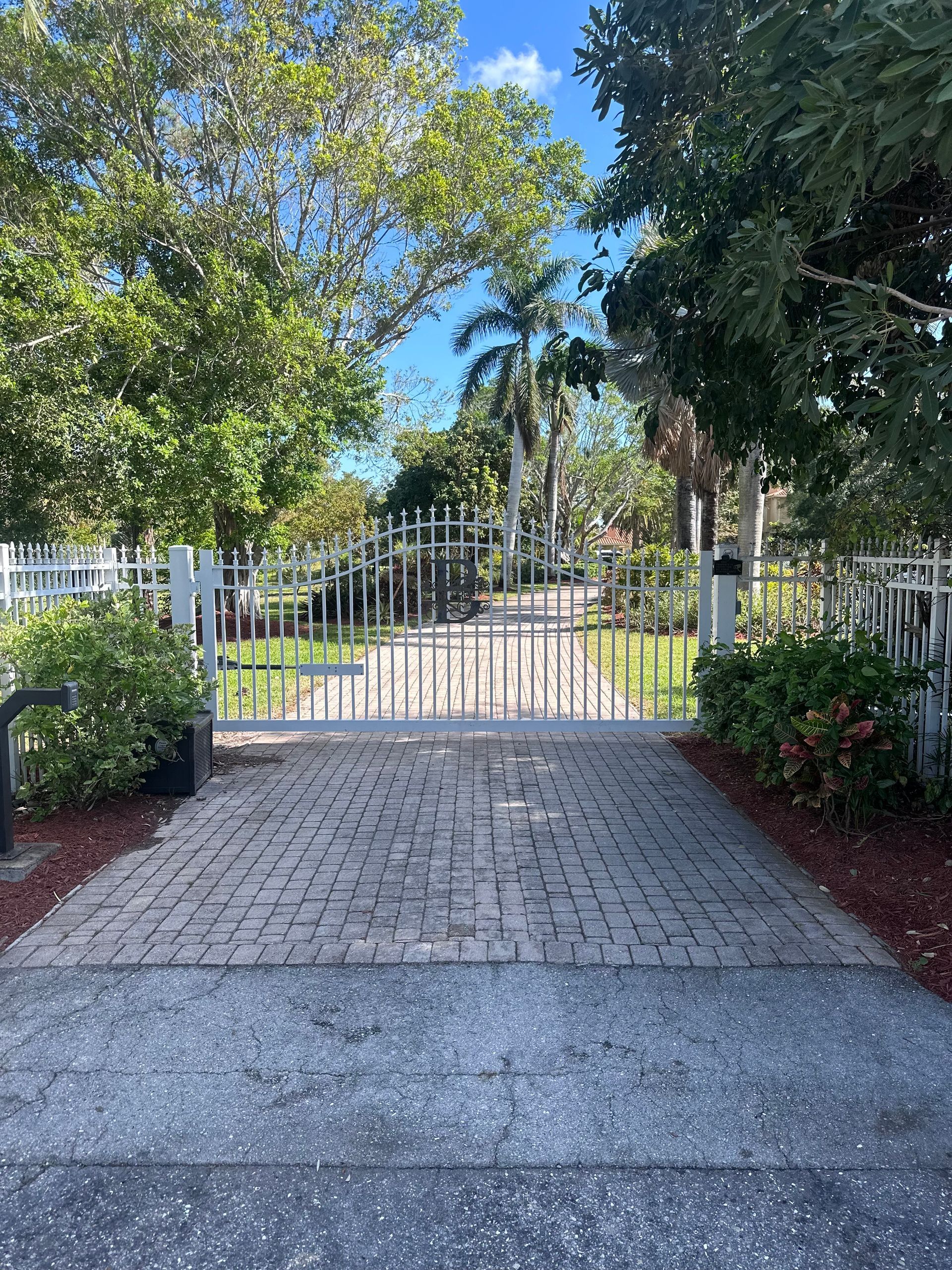 White gated entrance on a brick paved road, surrounded by lush green trees and bushes under a blue sky.