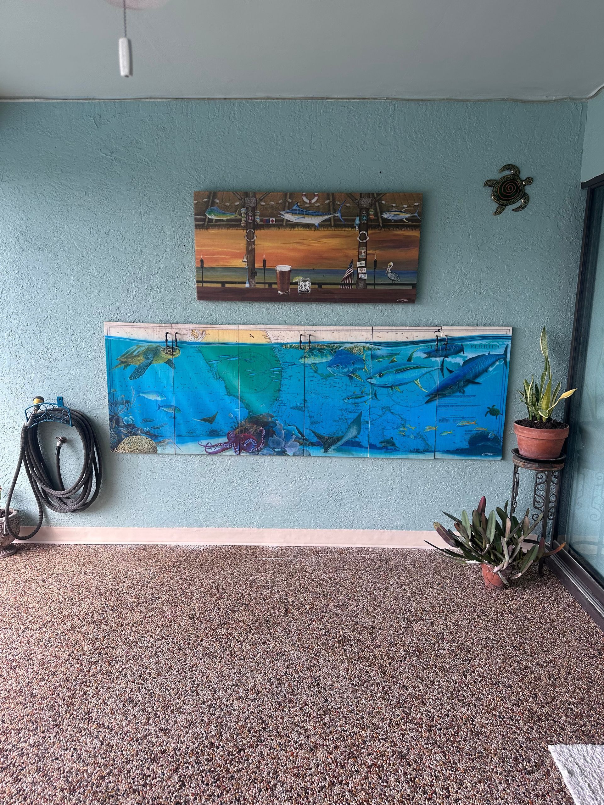 A room with an underwater painting, landscape art, and decorative plants; speckled floor and blue textured walls.