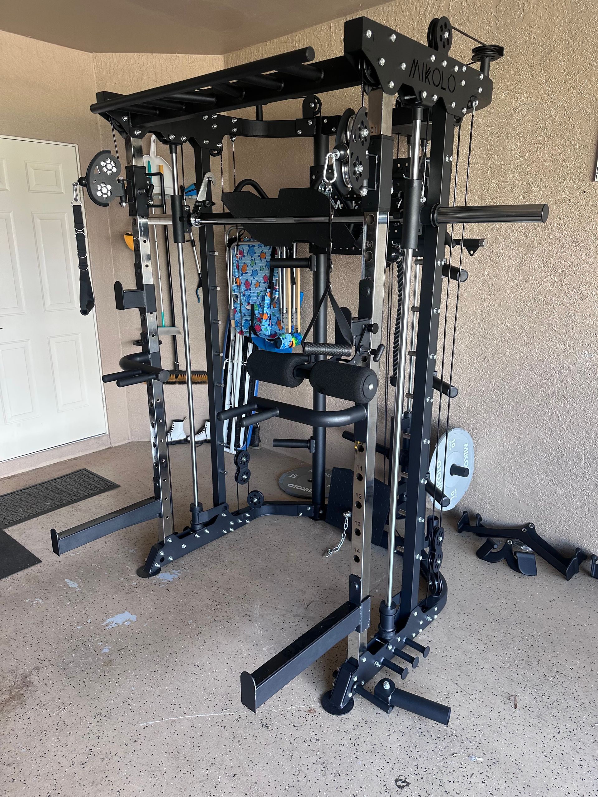 Black multi-gym fitness machine set up in a garage, featuring a smith machine, cable pulleys, and various attachments.