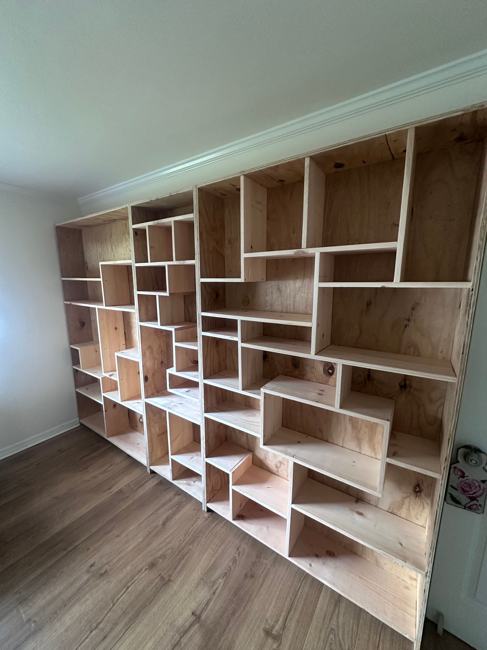 A large, built-in plywood bookcase with irregularly shaped compartments against a white wall and wood floor.