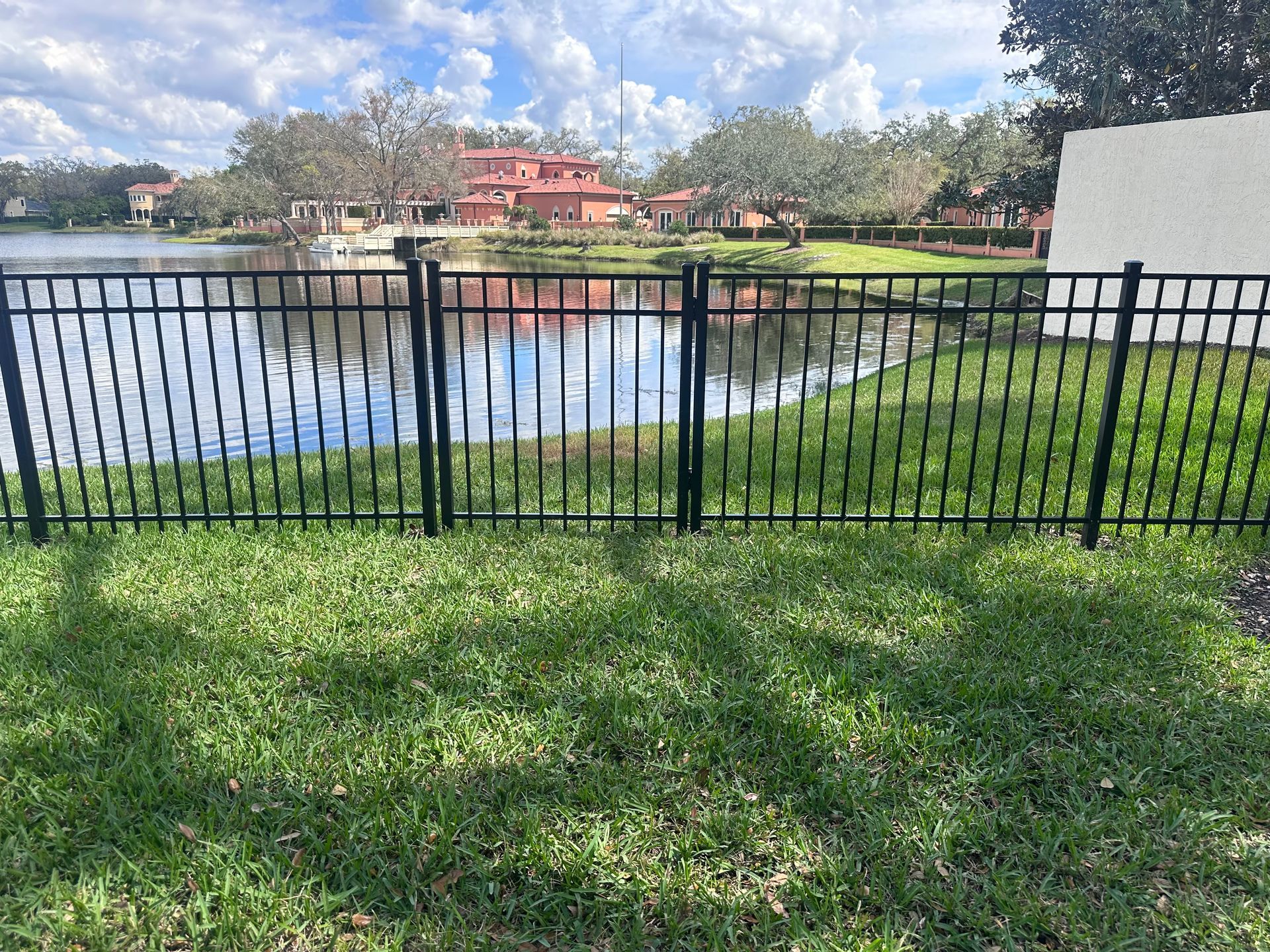 Black metal fence bordering green grass, lake, and distant buildings under a blue sky.