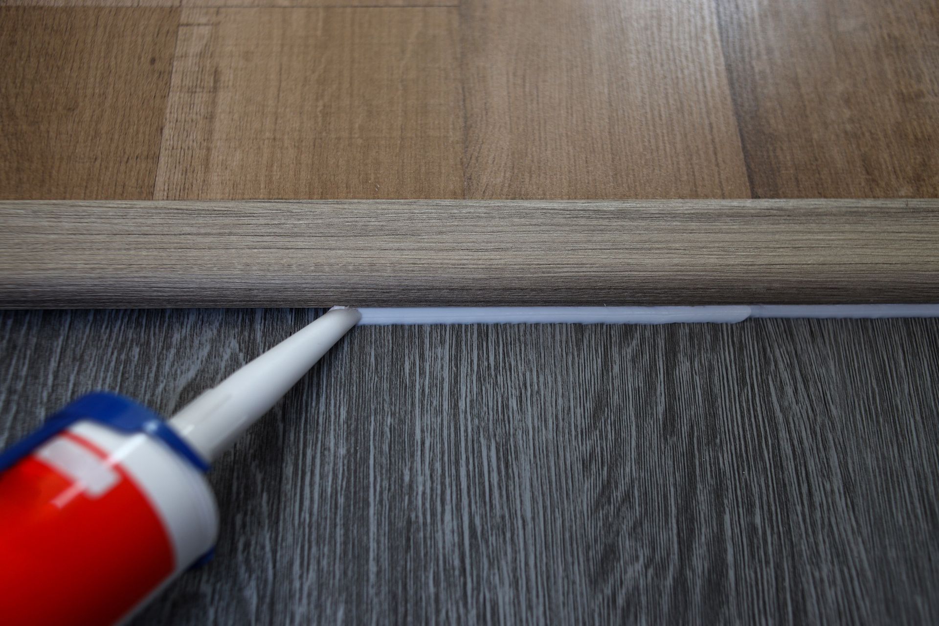 A caulk gun applies white sealant along the seam between a wooden floor and baseboard.