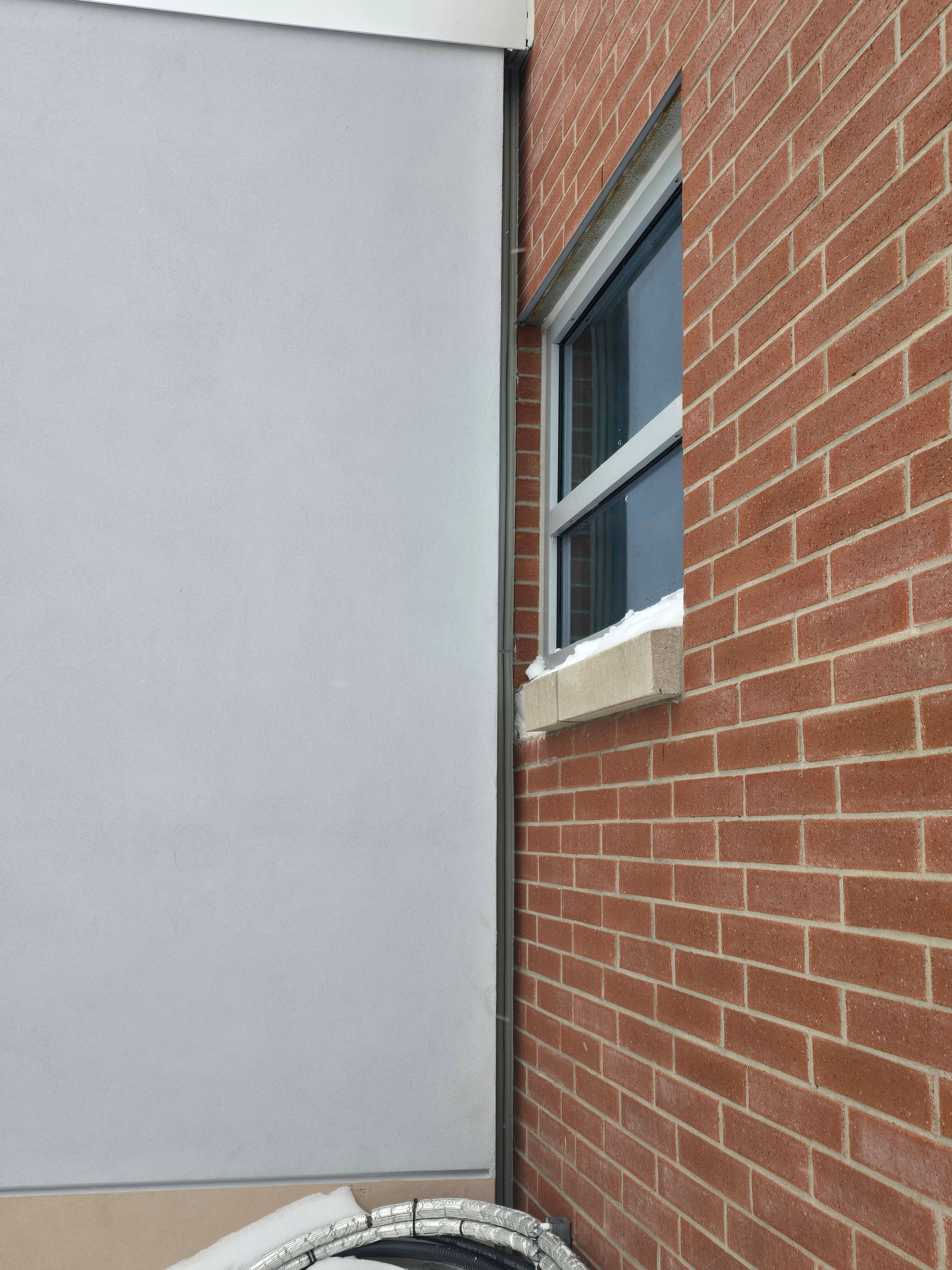 Narrow gap between a gray wall and a red brick building with a window and snow along the base