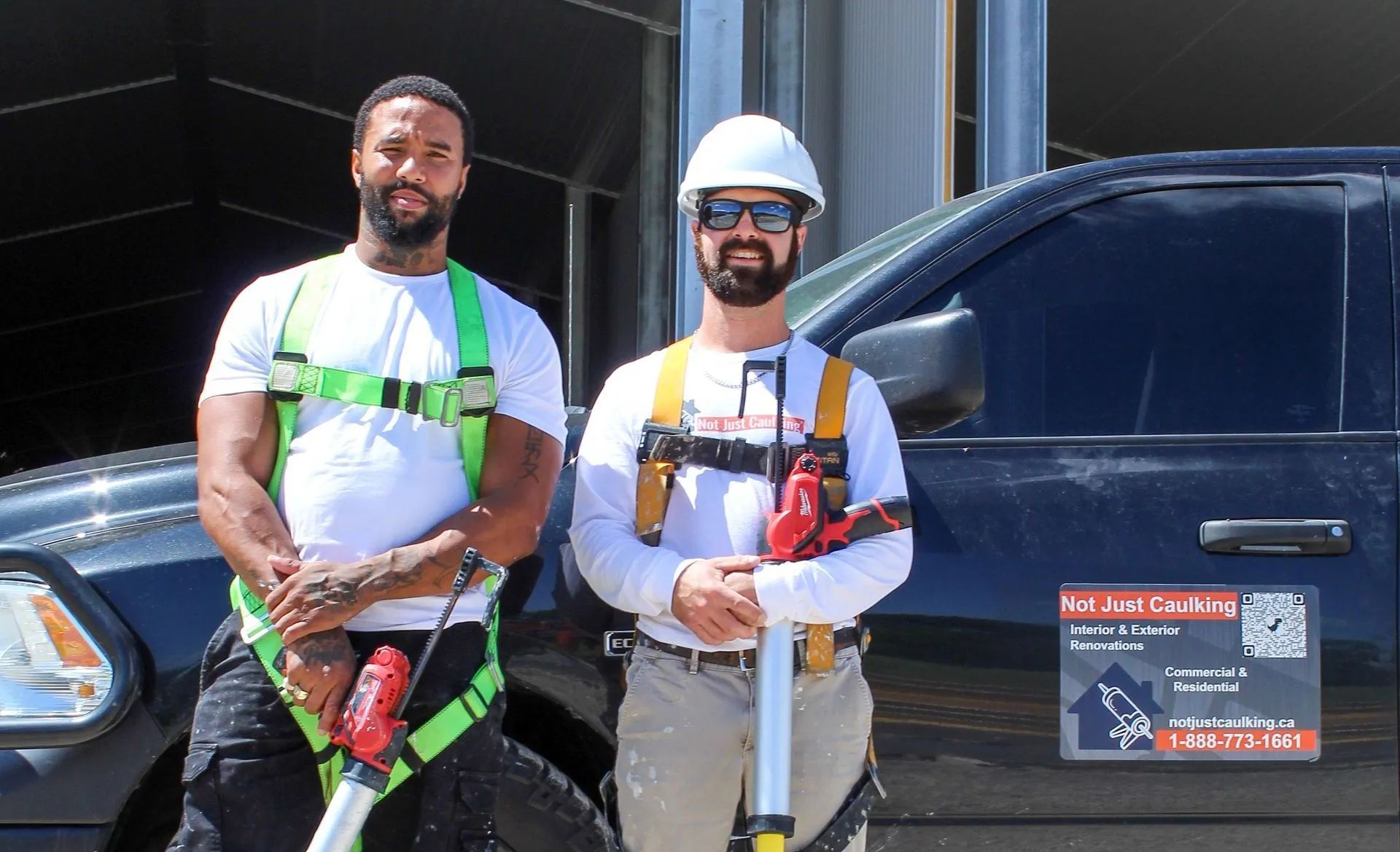 Two workers in safety gear stand beside a black vehicle outdoors, holding tools and hoses.