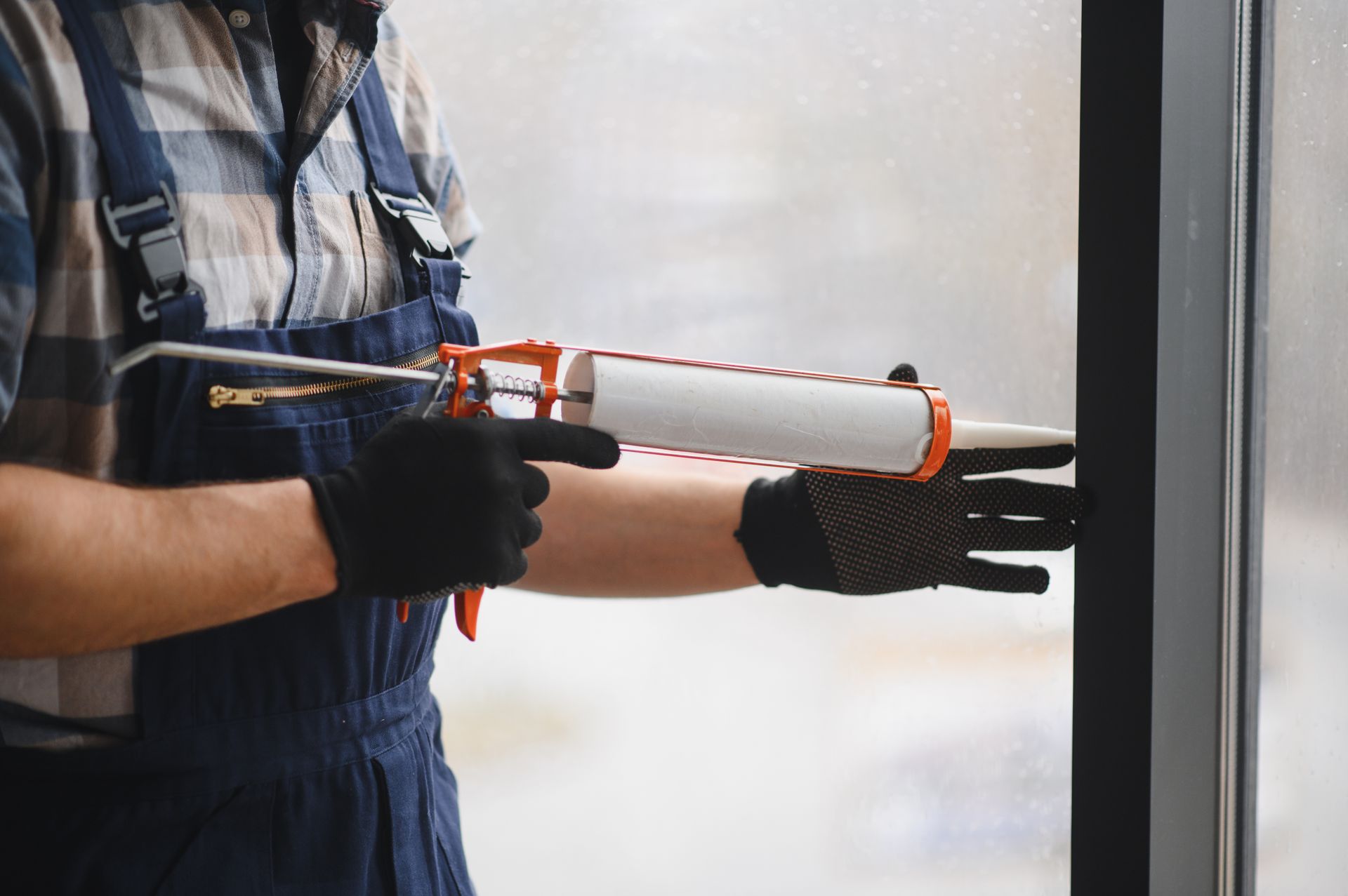 Person wearing overalls and gloves, using a caulk gun to seal a window frame.