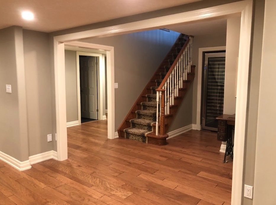 Wooden-floored interior with stairway, entryway, and doorway. Gray walls, white trim, and a dark carpeted staircase.