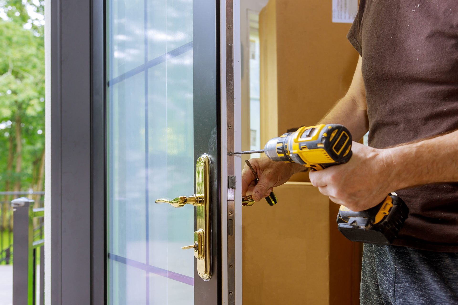 Person using a yellow drill to install door hardware on a glass-paned door outdoors.