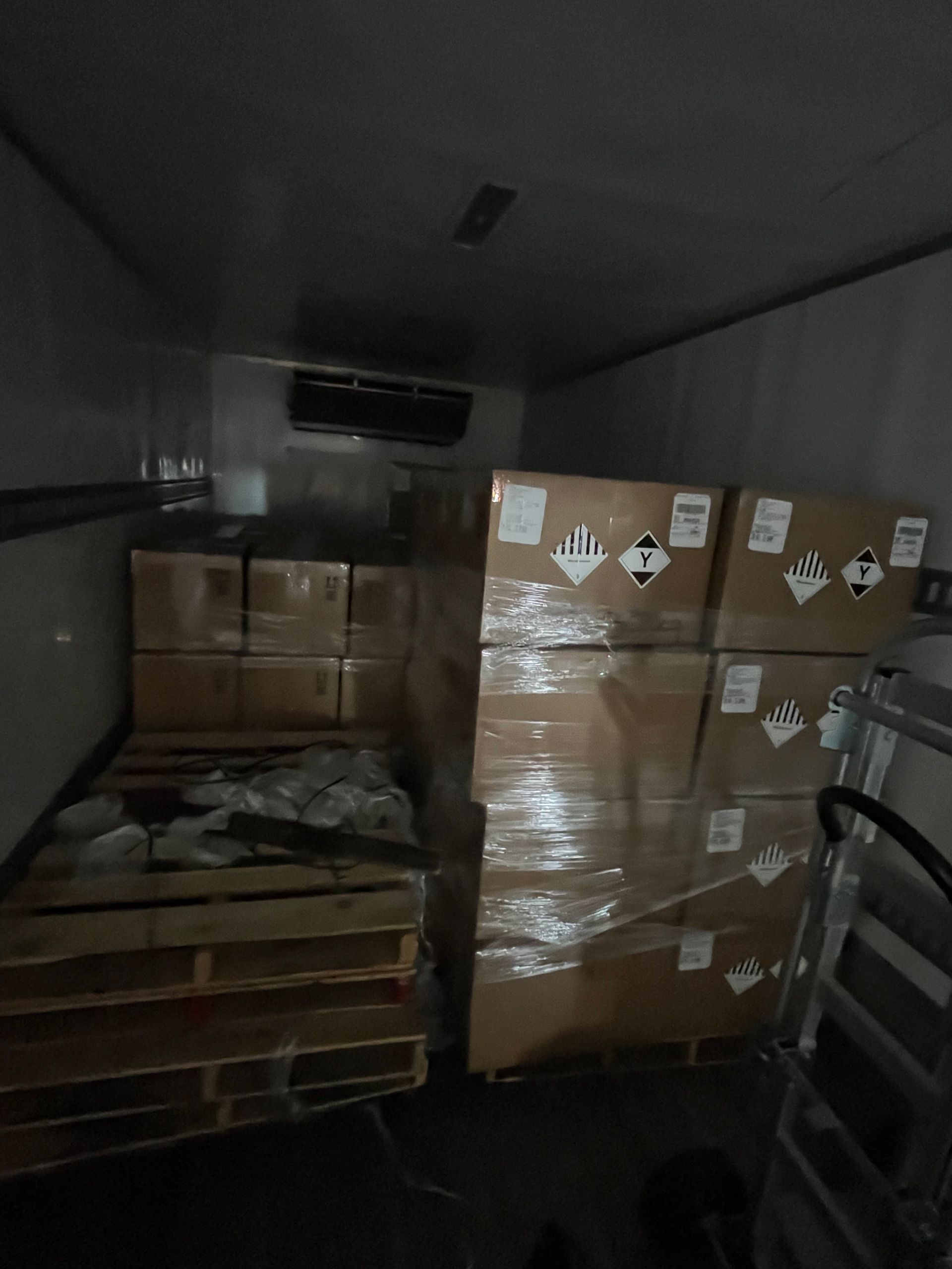 Inside a refrigerated cargo container: boxes stacked on pallets, wrapped in plastic, with hazardous material labels.