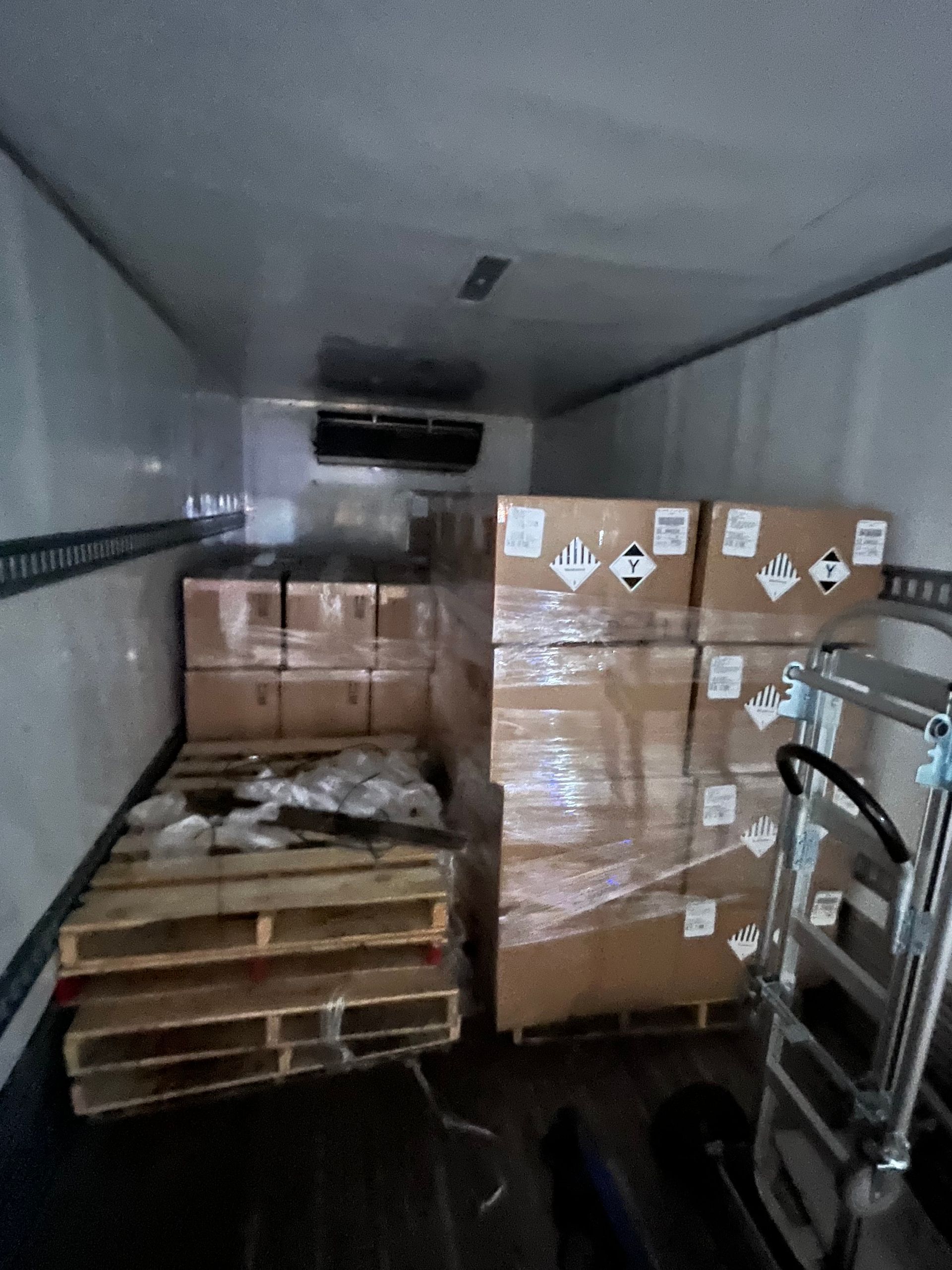 Inside a refrigerated truck, boxes of chemicals on pallets and a hand truck.