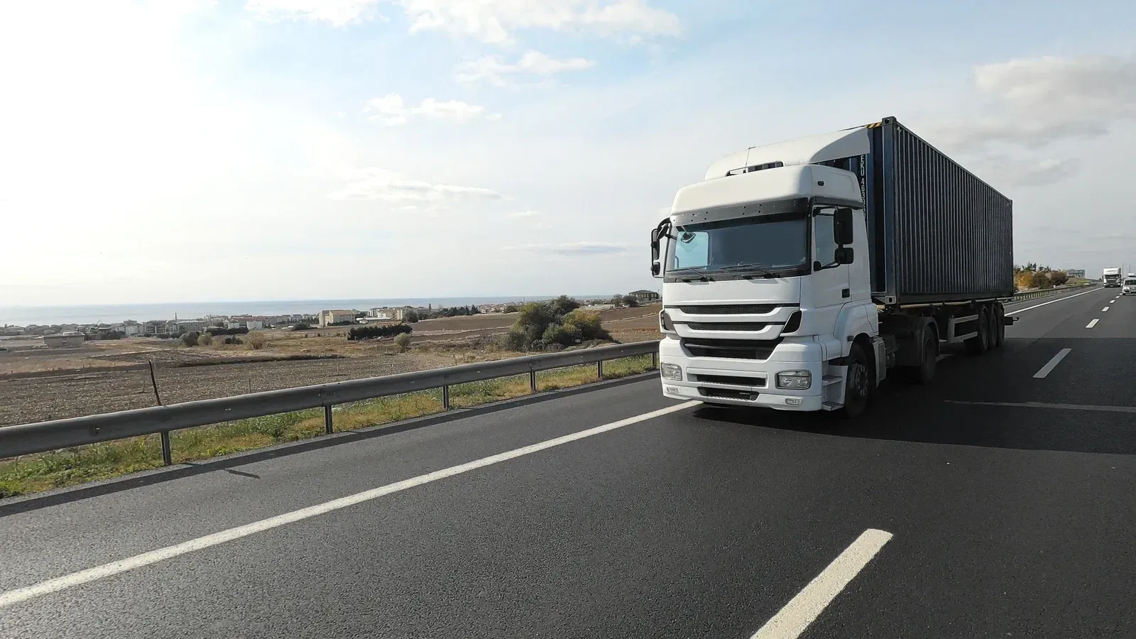 White semi-truck driving on a highway, roadside view with ocean and buildings in the background.