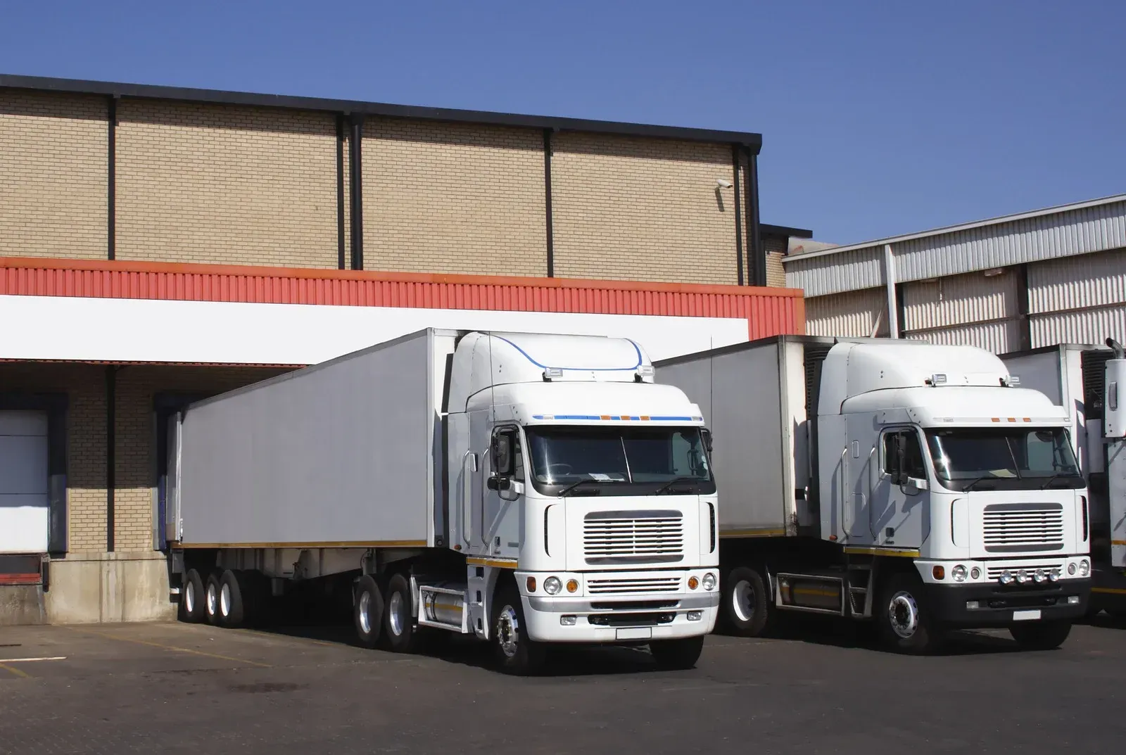 Two white semi-trucks parked at a loading dock, near a brick warehouse.