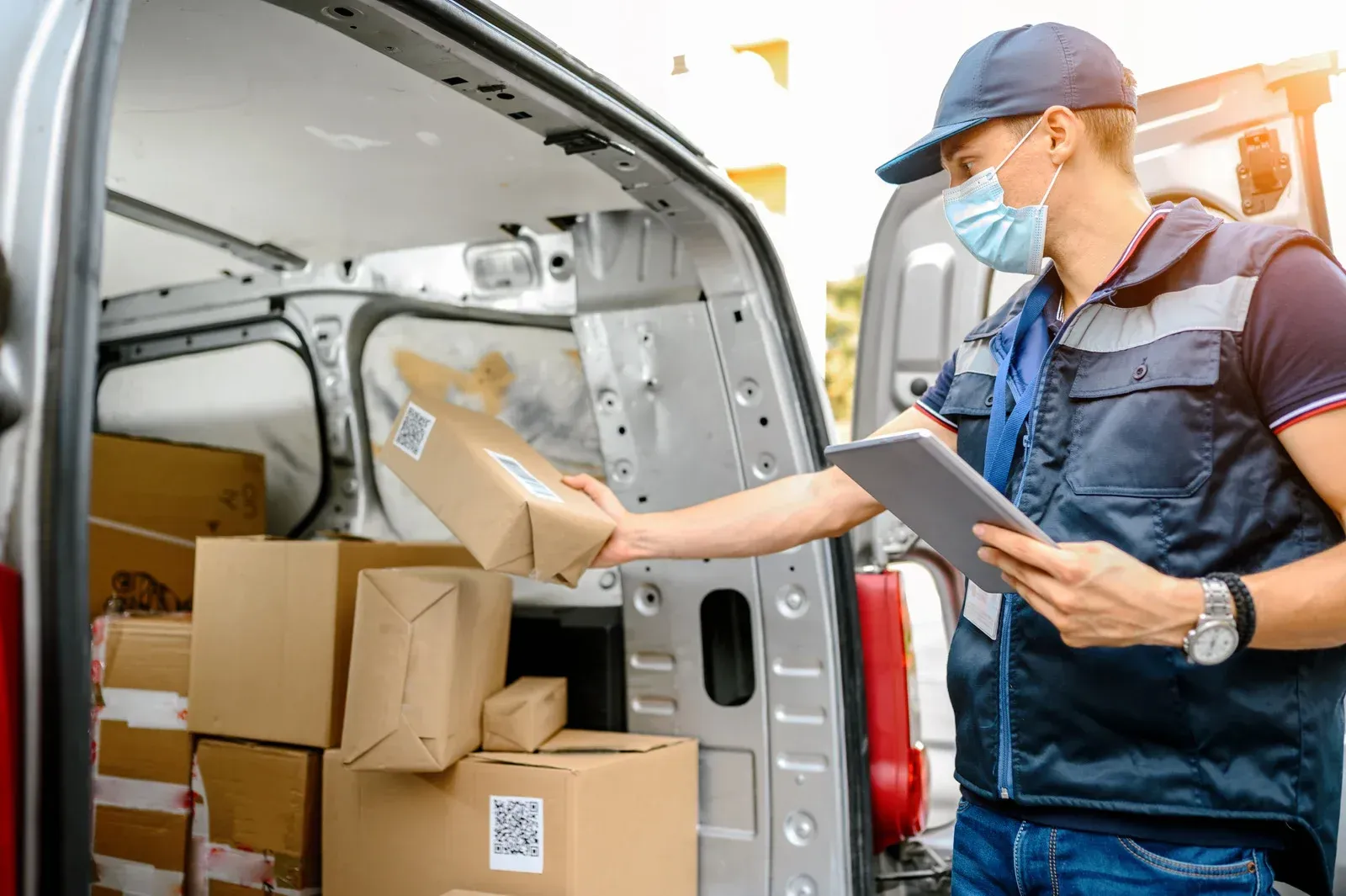 Delivery person wearing mask unloading packages from a van; sunlight.