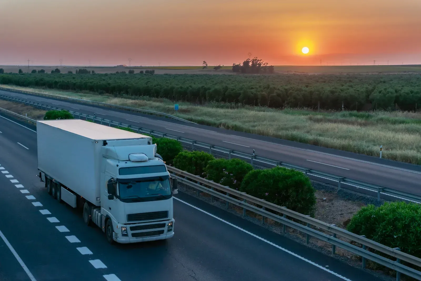 Semi-truck driving on highway at sunset, passing green fields.