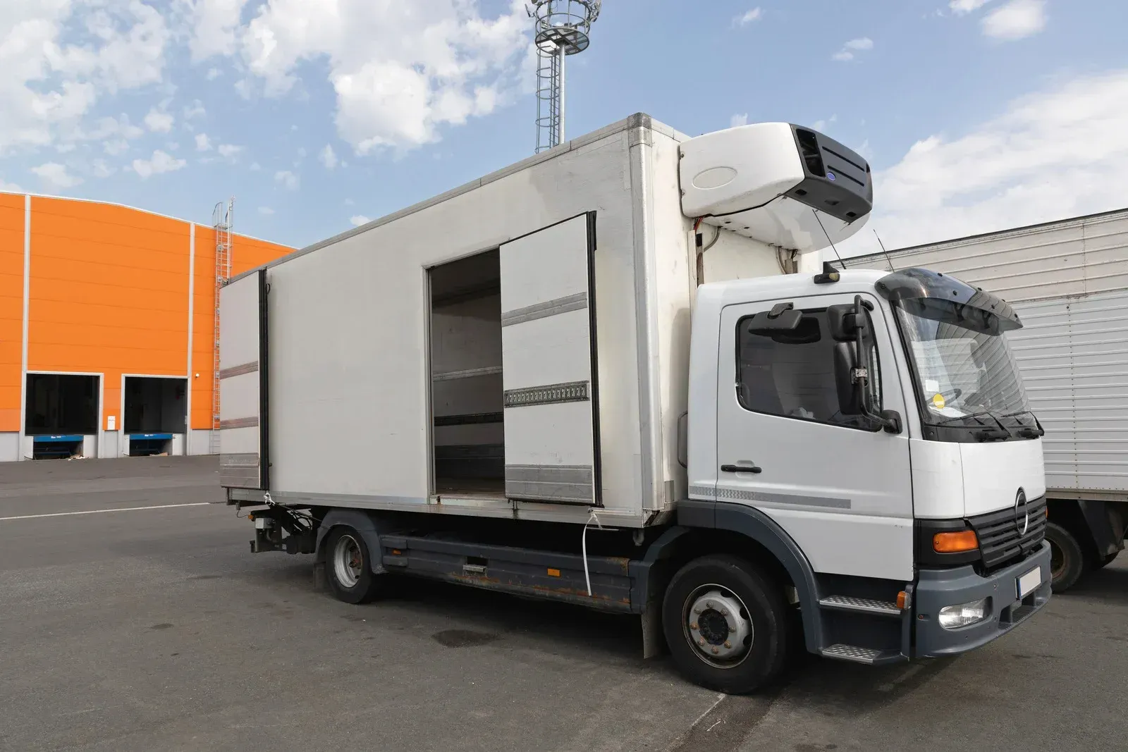 White refrigerated delivery truck with open side doors parked outside a warehouse.