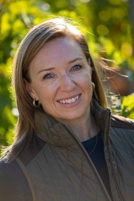 Woman smiling, wearing a khaki vest, with blonde hair, in an outdoor setting with green foliage.