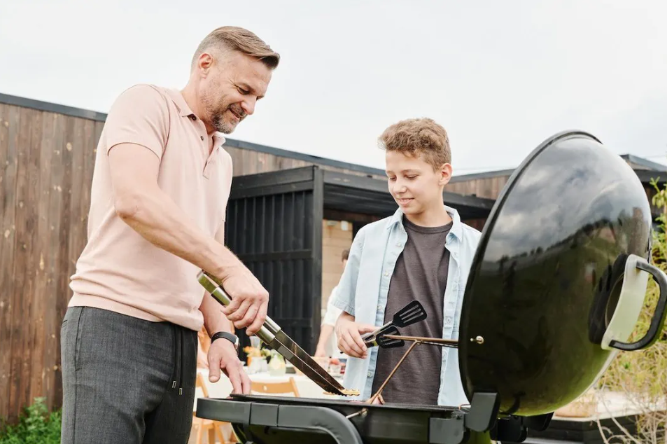 Man and teen boy grilling food outdoors, smiling; gray pants, pink shirt, black grill.