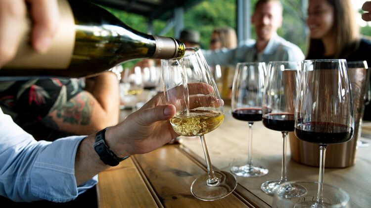 Person pouring white wine into a glass at a table with other wine glasses and people in the background.