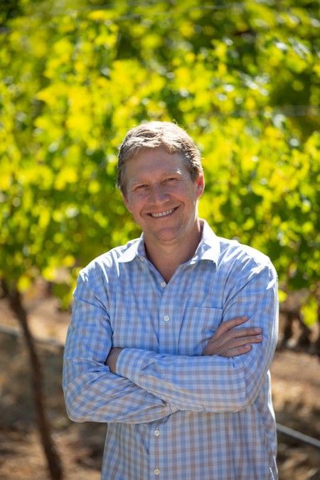 Man smiling with arms crossed, standing outdoors in front of a vineyard, wearing a blue checkered shirt.