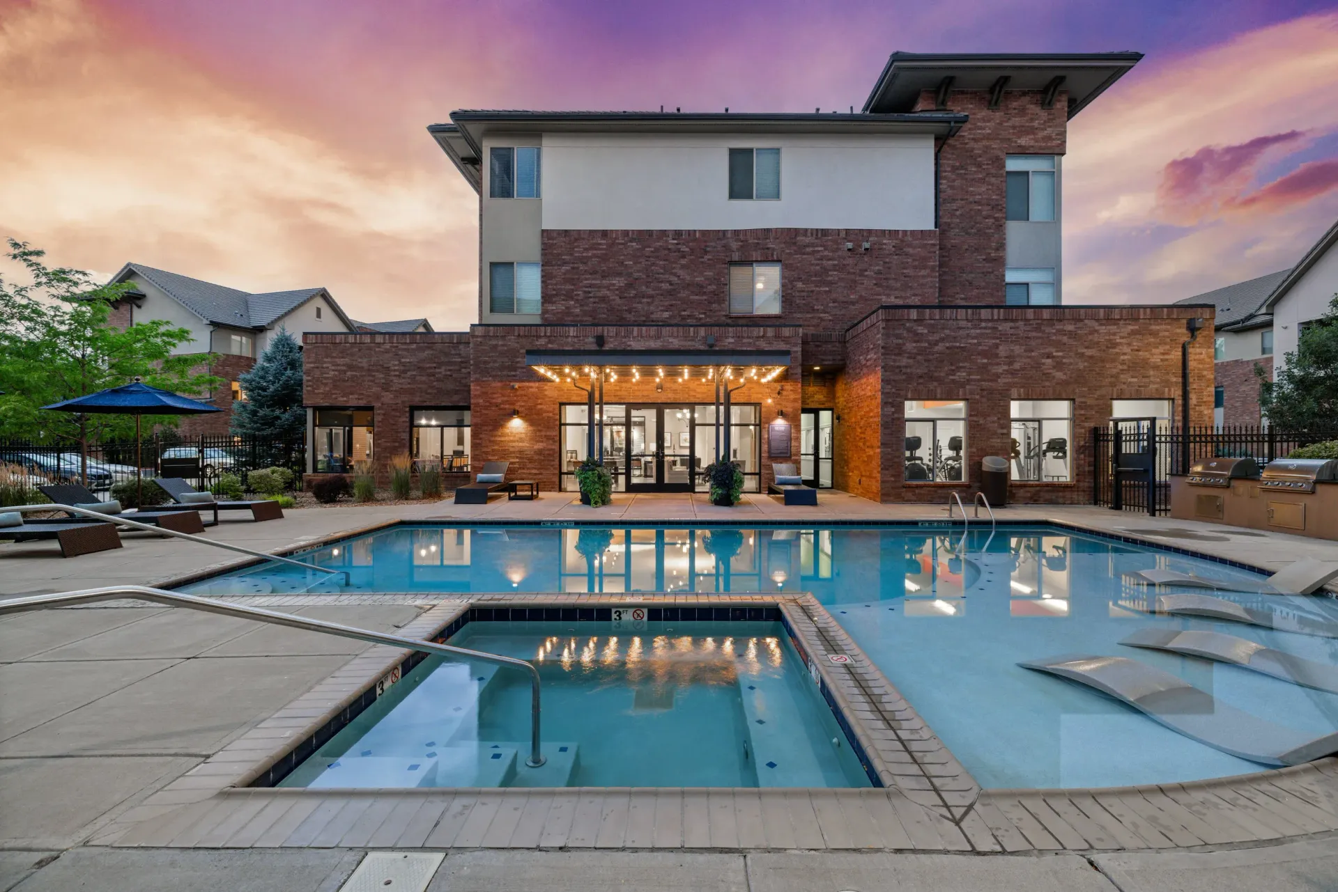 Exterior view of a brick apartment pool area with lounge chairs at sunset.